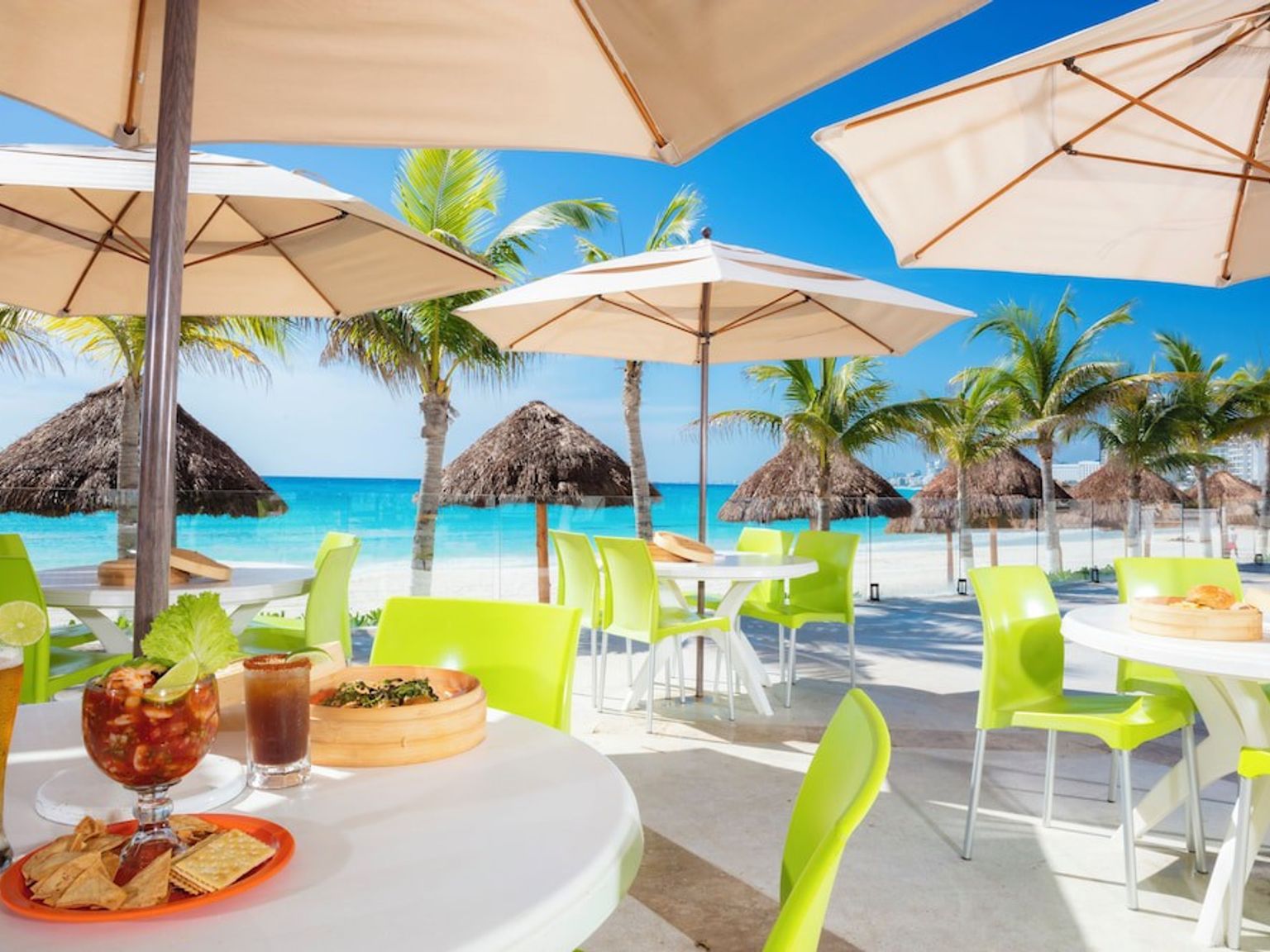 Utomhus strandrestaurang med mat och dryck, limegröna stolar, parasoller, palmer och en turkos ocean.