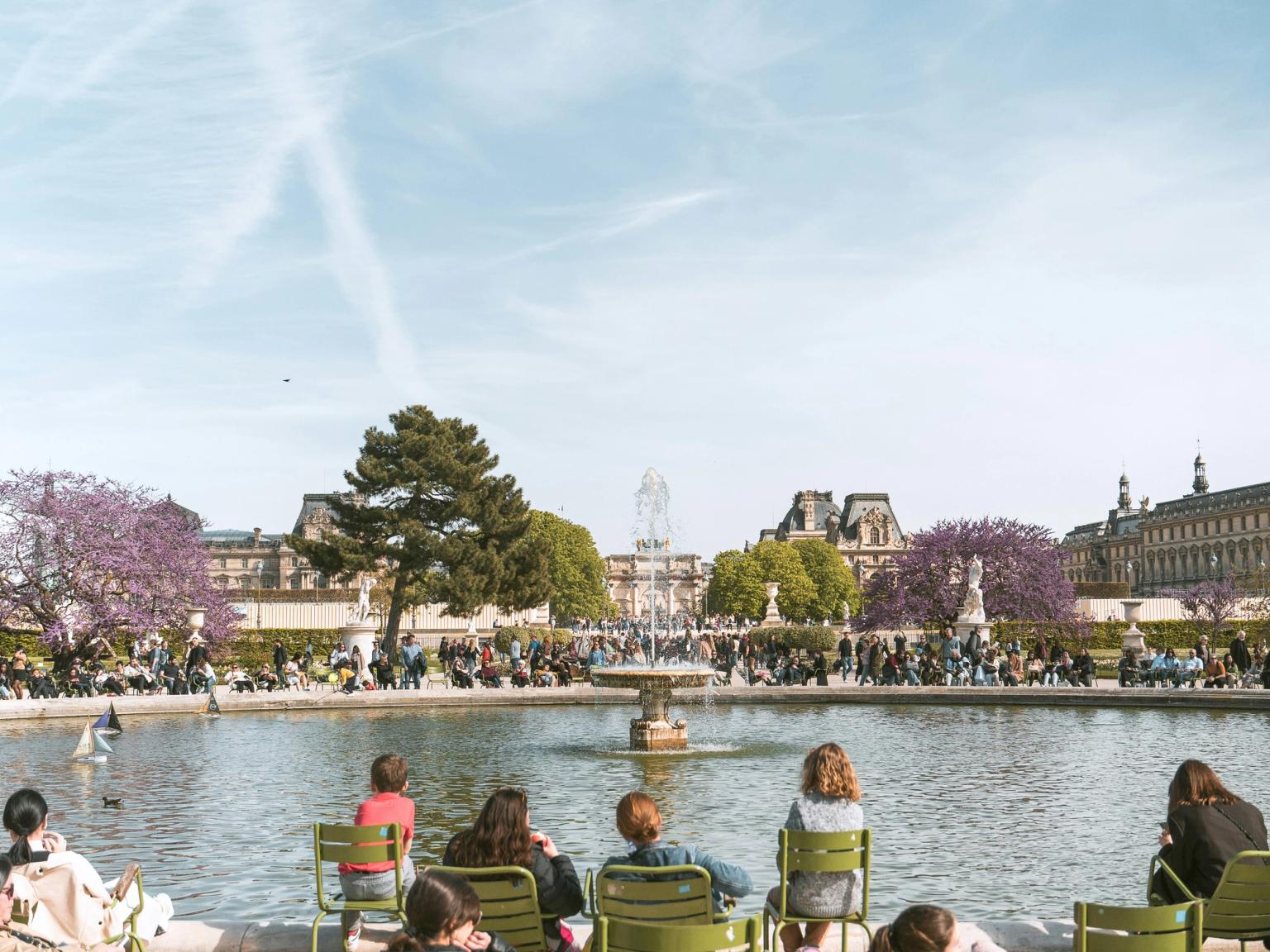 People relaxing in green chairs facing a pond with fountains, surrounded by trees and historic buildings under a sunny, contrail-filled sky. Paris.