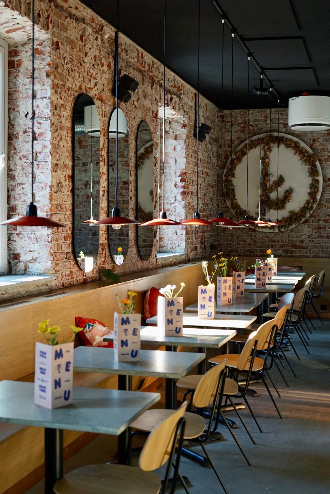 Et moderne restaurantinteriør med blotlagte murstensvægge, en lang bænk, lyse borde, træstole og røde pendellamper. Hvert bord har en menu, der holder en lille blomsterdekoration.