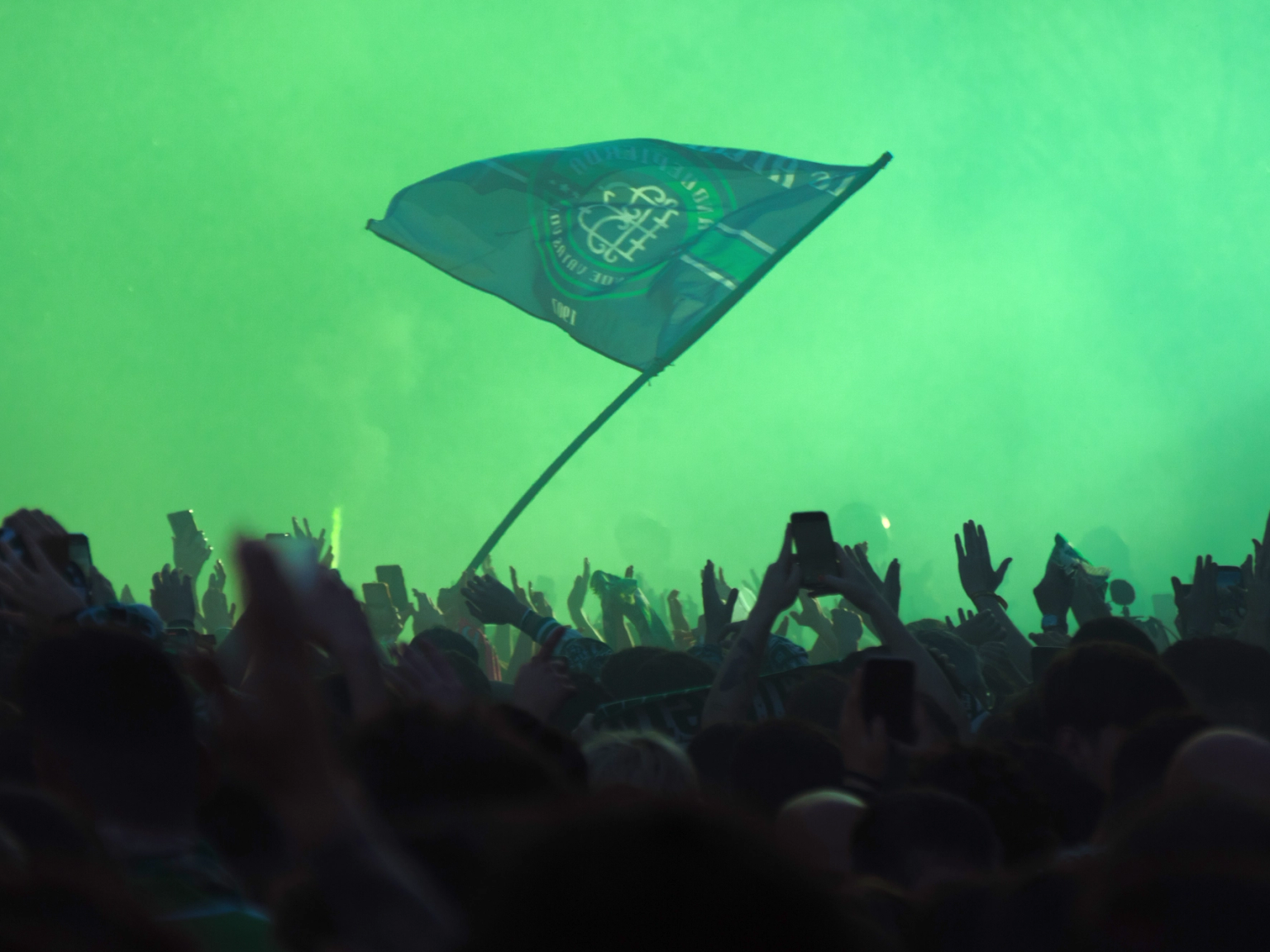 A crowd of people with raised hands and phones, illuminated by green light, with a green flag waving above them.