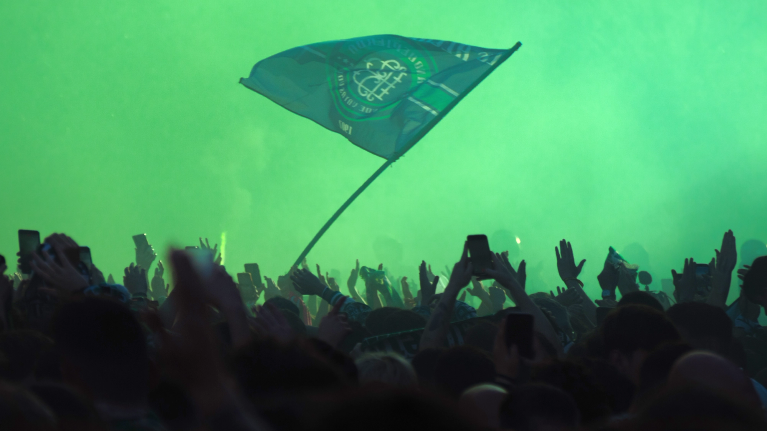 A crowd of people with raised hands and phones, illuminated by green light, with a green flag waving above them.