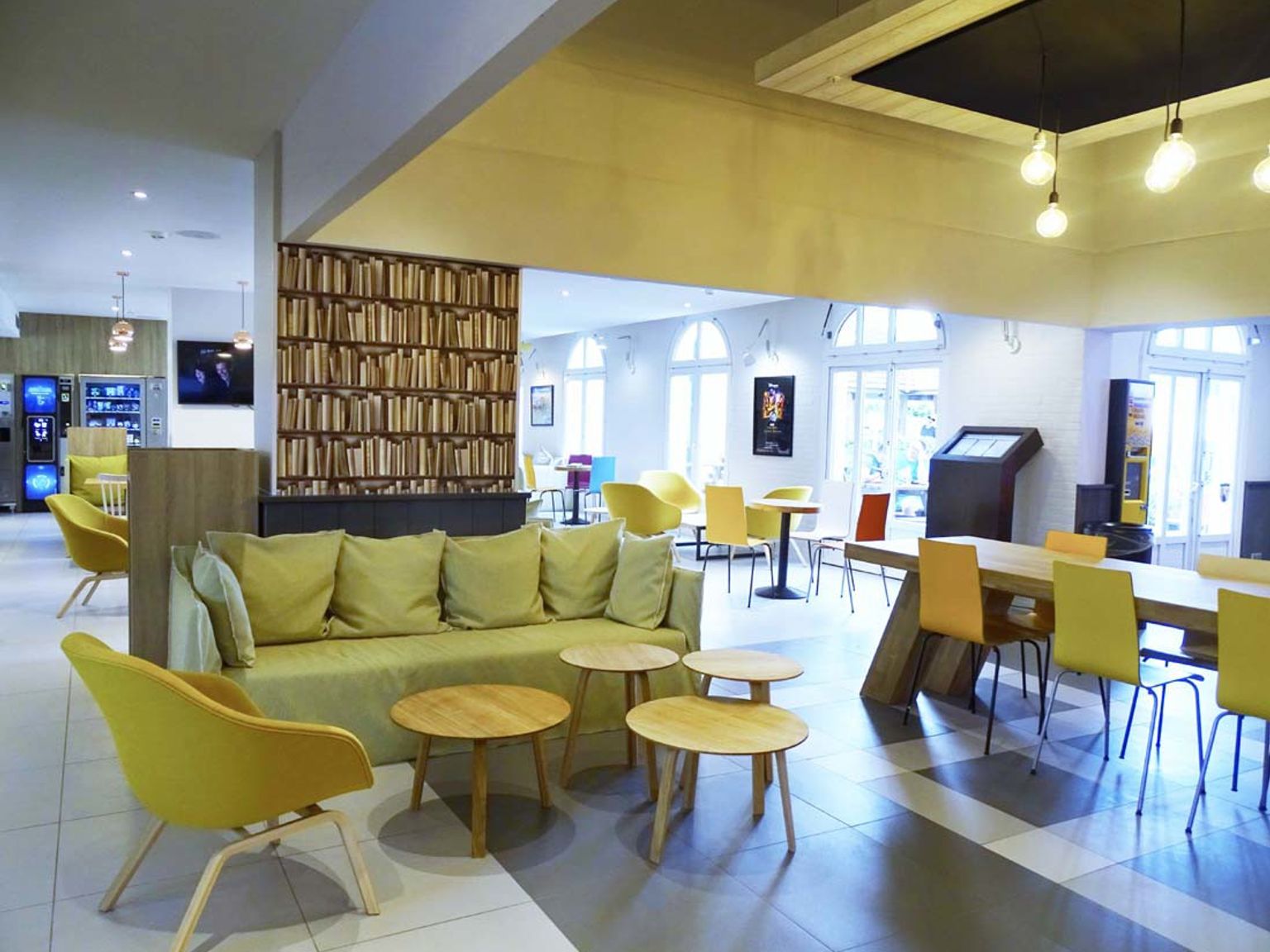 En ljus, modern lounge med gula soffor, träbord, en bokhyllsmönstrad vägg och en matplats med färgglada stolar. Hotel Campanile Val de France vid Disneyland® Paris