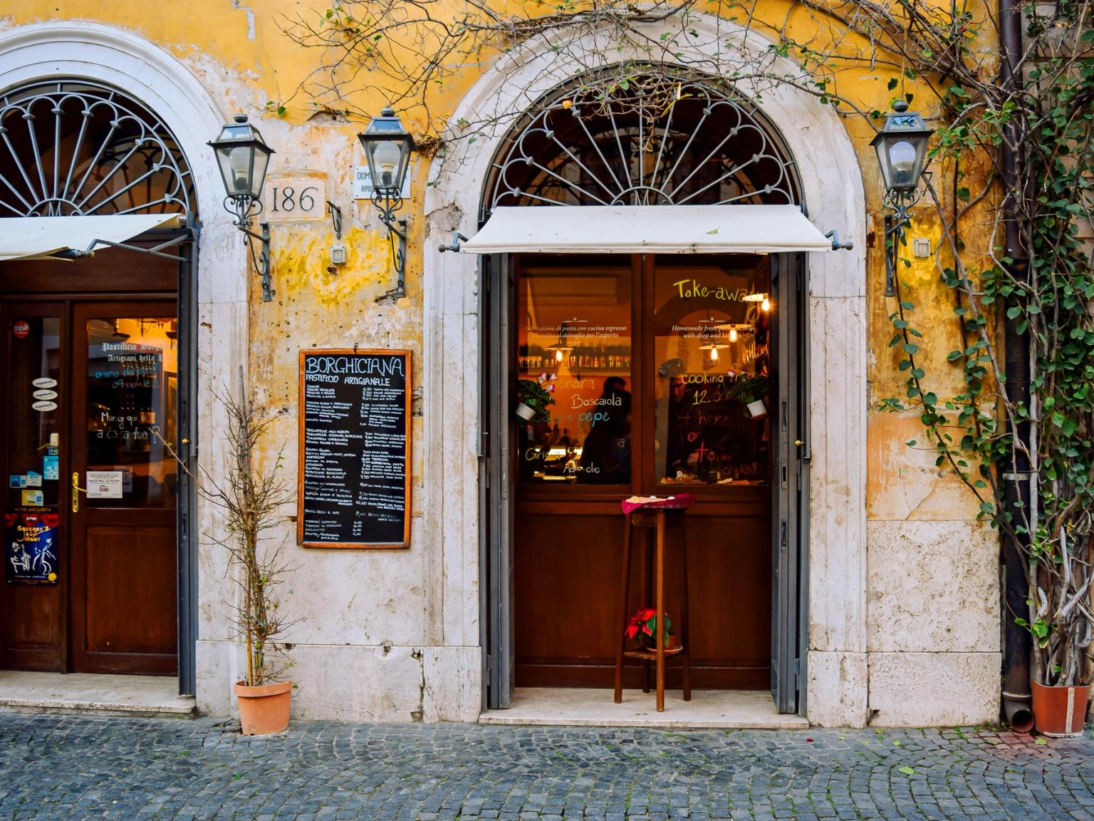 Dwoje łukowatych drewnianych drzwi rustykalnego budynku w Rzymie z tablicą menu i pnącymi winoroślami.