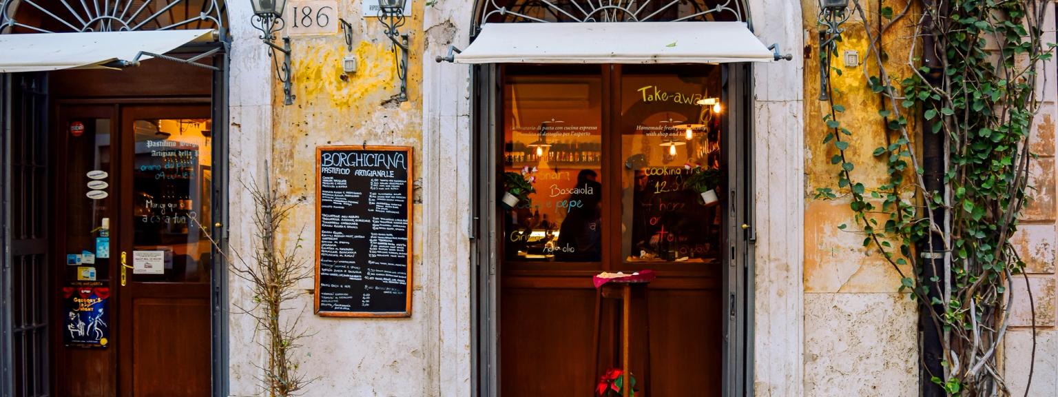 Två välvda trädörrar på en rustik byggnad i Rom med en menytavla och klättrande vinrankor.