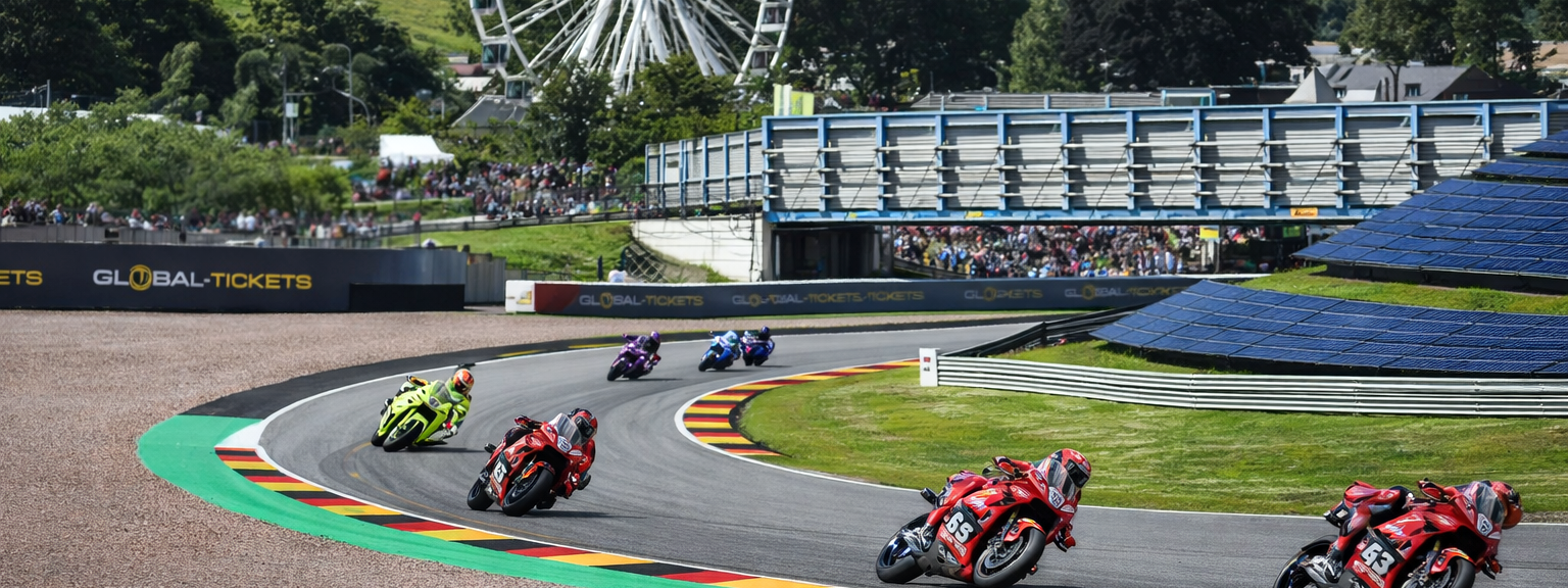 Motorcycles race on a track with German flag curbs, a Ferris wheel, and spectators.