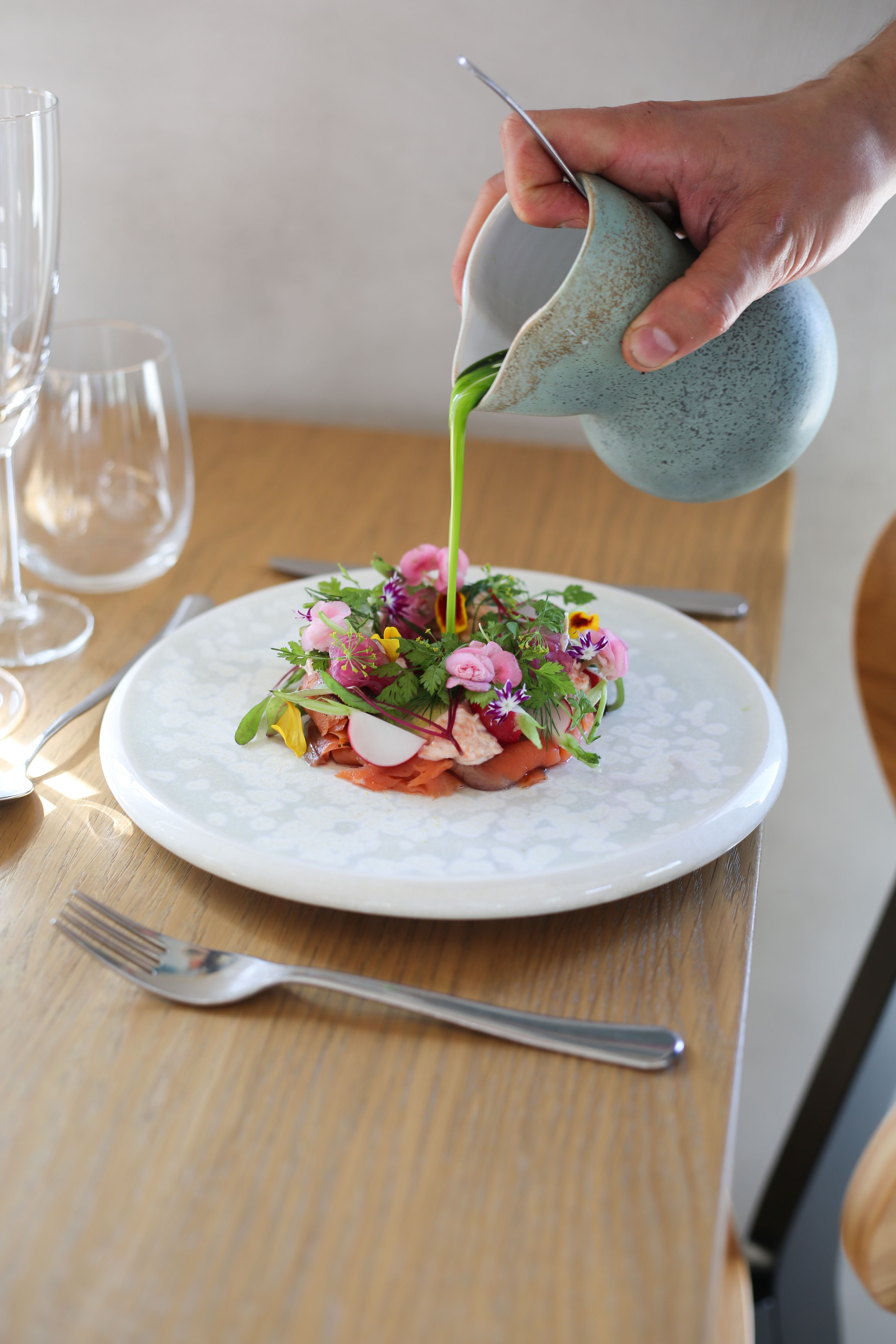 En hånd hælder en livlig grøn sauce over en kunstfærdigt anrettet ret med spiselige blomster, urter og skåret protein på en hvid tallerken, der står på et træbord.
