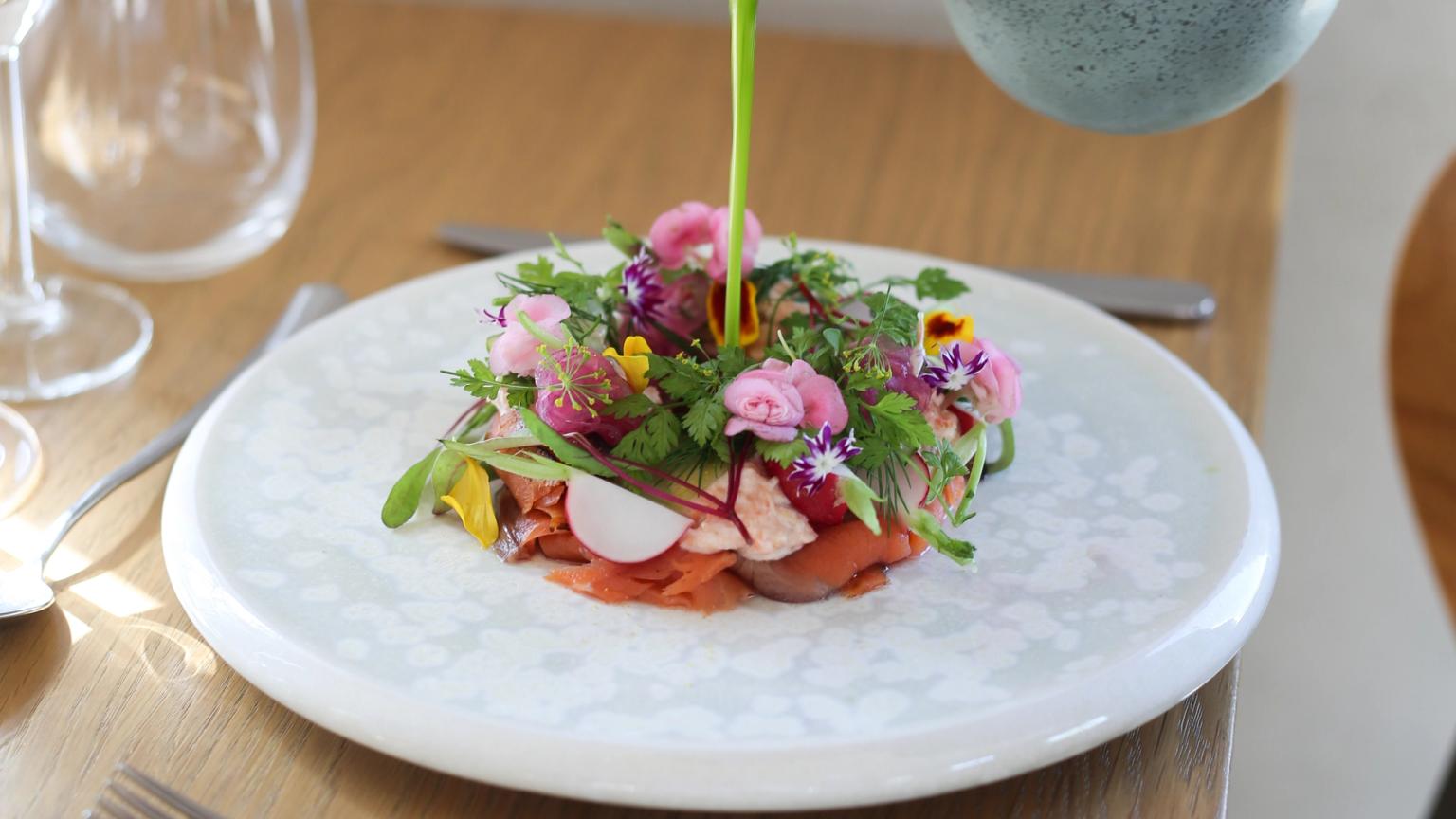 En hånd hælder en livlig grøn sauce over en kunstfærdigt anrettet ret med spiselige blomster, urter og skåret protein på en hvid tallerken, der står på et træbord.