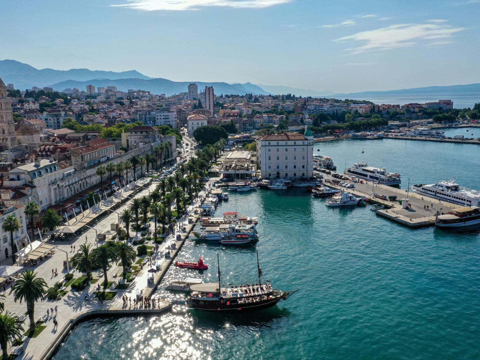 Flyfoto av en livlig kystby med en palmekledd promenade, en travel havn, historisk arkitektur og fjell i bakgrunnen. Split, Kroatia.
