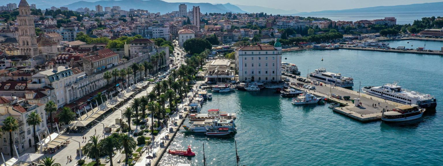 Flygvy över en livlig kuststad med en palmkantad strandpromenad, en trafikerad hamn, historisk arkitektur och berg i bakgrunden. Split, Kroatien.