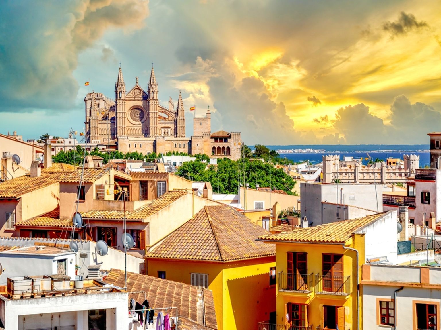 Die Kathedrale La Seu mit Blick auf die Terrakottadächer von Palma de Mallorca und das Meer unter einem dramatischen goldenen Himmel.