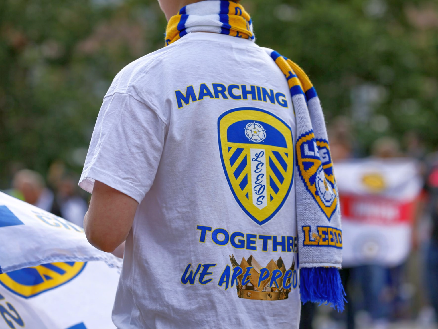 Costas de uma pessoa vestindo uma camiseta branca do Leeds United e cachecol, com "MARCHING TOGETHER WE ARE PROUD" na camisa.