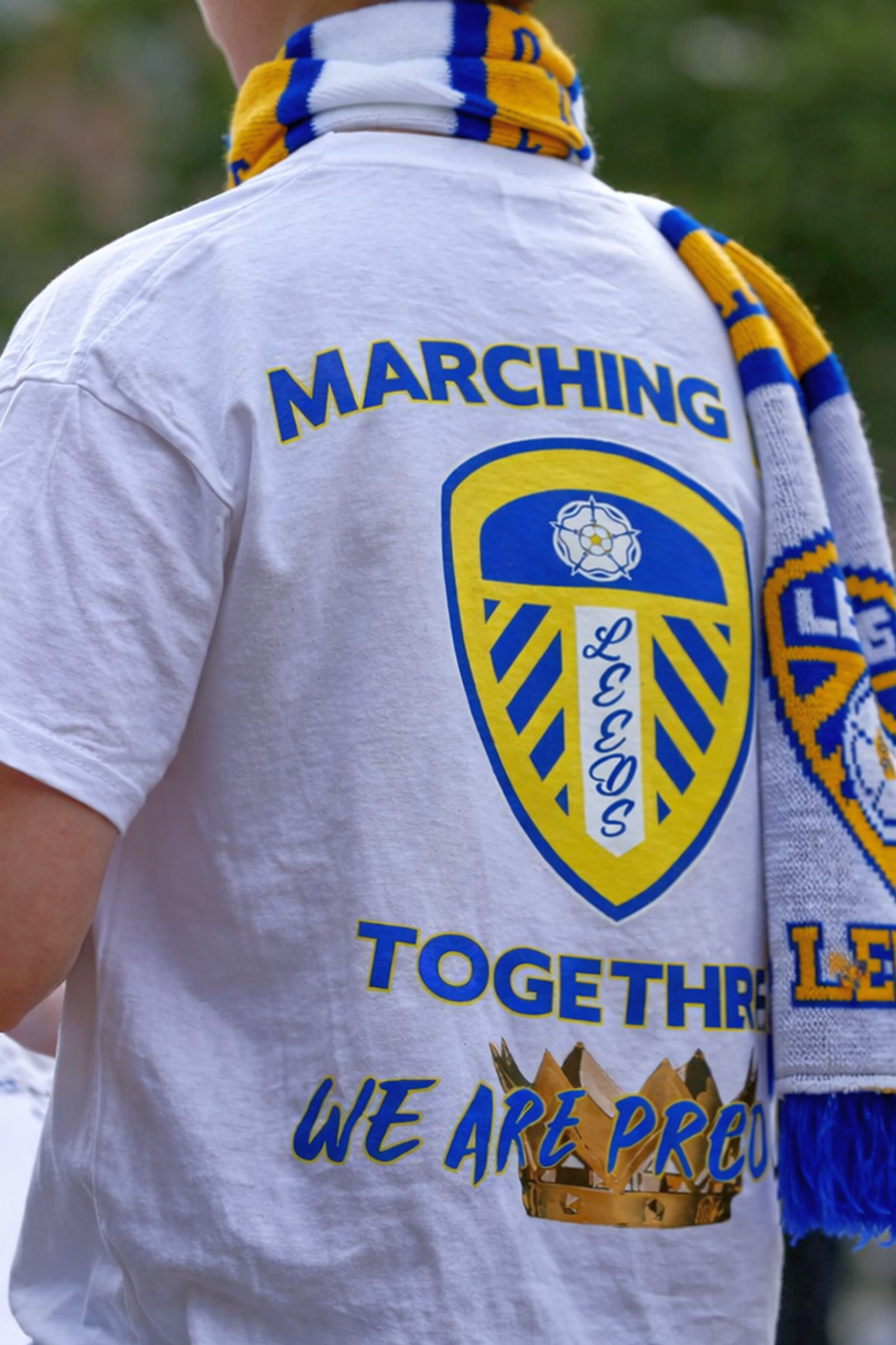 Costas de uma pessoa vestindo uma camiseta branca do Leeds United e cachecol, com "MARCHING TOGETHER WE ARE PROUD" na camisa.
