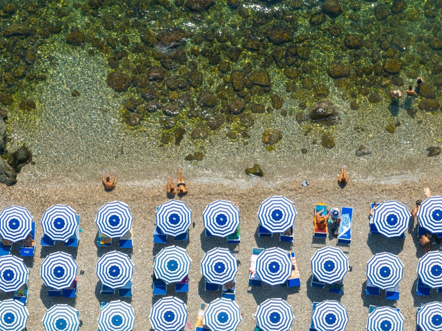 Fugleperspektiv av en strand med rekker av blå- og hvitstripete parasoller, solbadere og klart vann med steiner og svømmere.