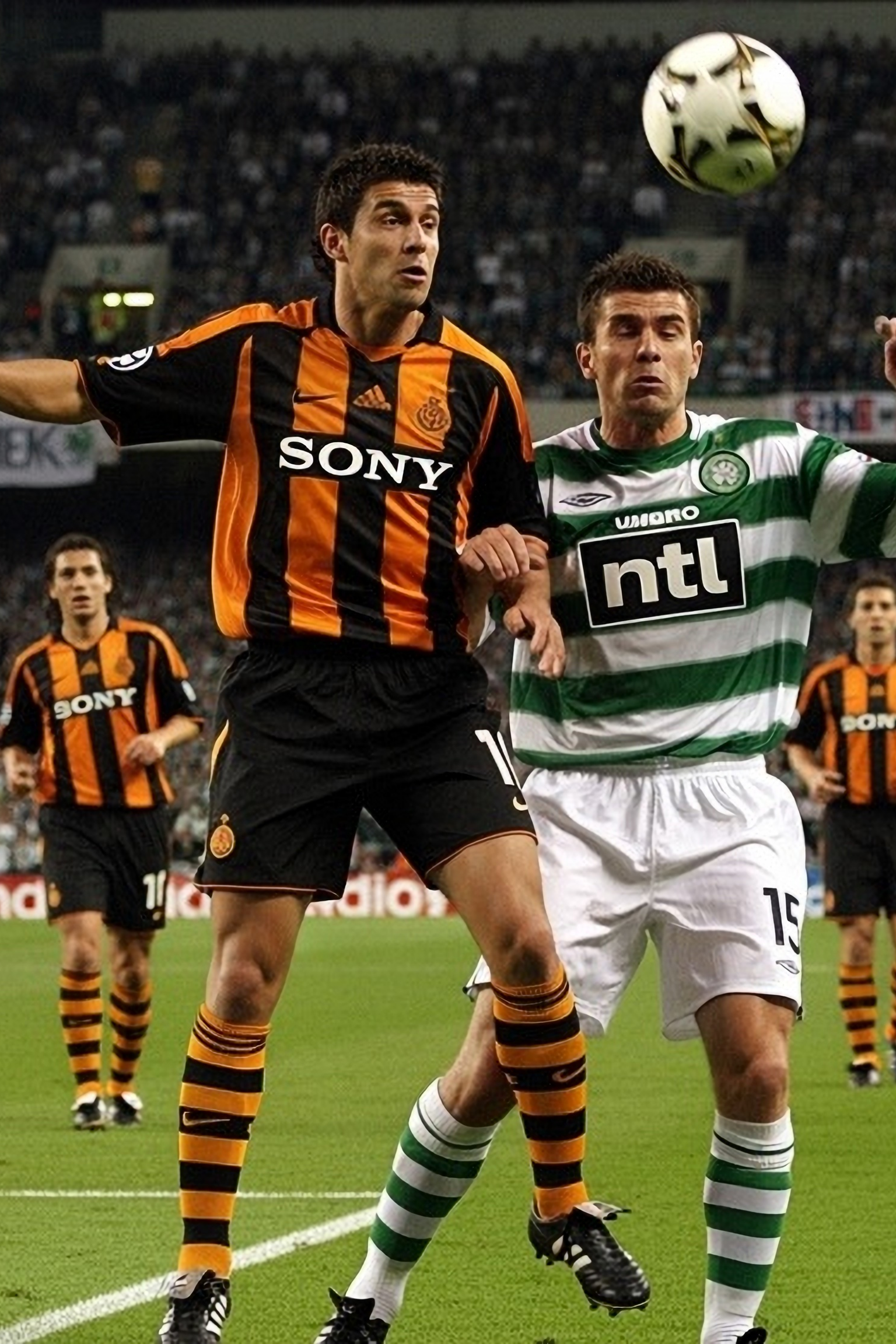 Två fotbollsspelare, en i orange och svart, en i grönt och vitt, kämpar om en luftboll.
