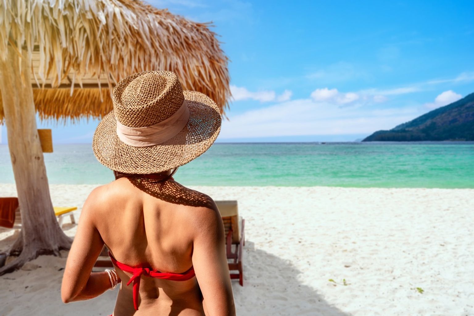 Rückansicht einer Person mit Strohhut und rotem Bikini, die auf einen tropischen Strand mit türkisfarbenem Wasser blickt.