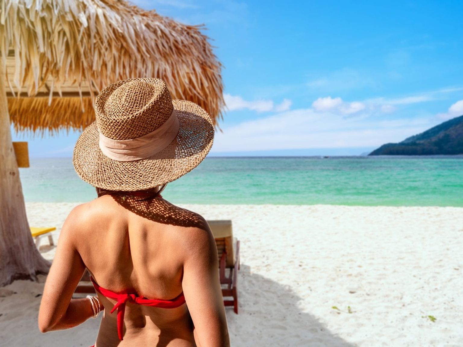 Bakfra av en person med stråhatt og rød bikini som ser ut over en tropisk strand med turkis vann.