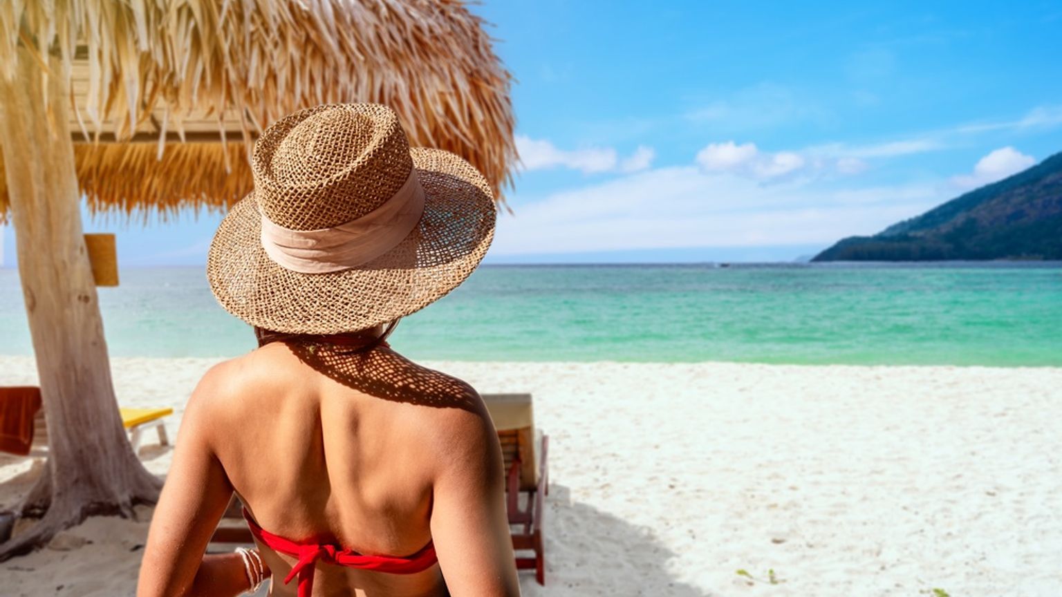 Bakfra av en person med stråhatt og rød bikini som ser ut over en tropisk strand med turkis vann.
