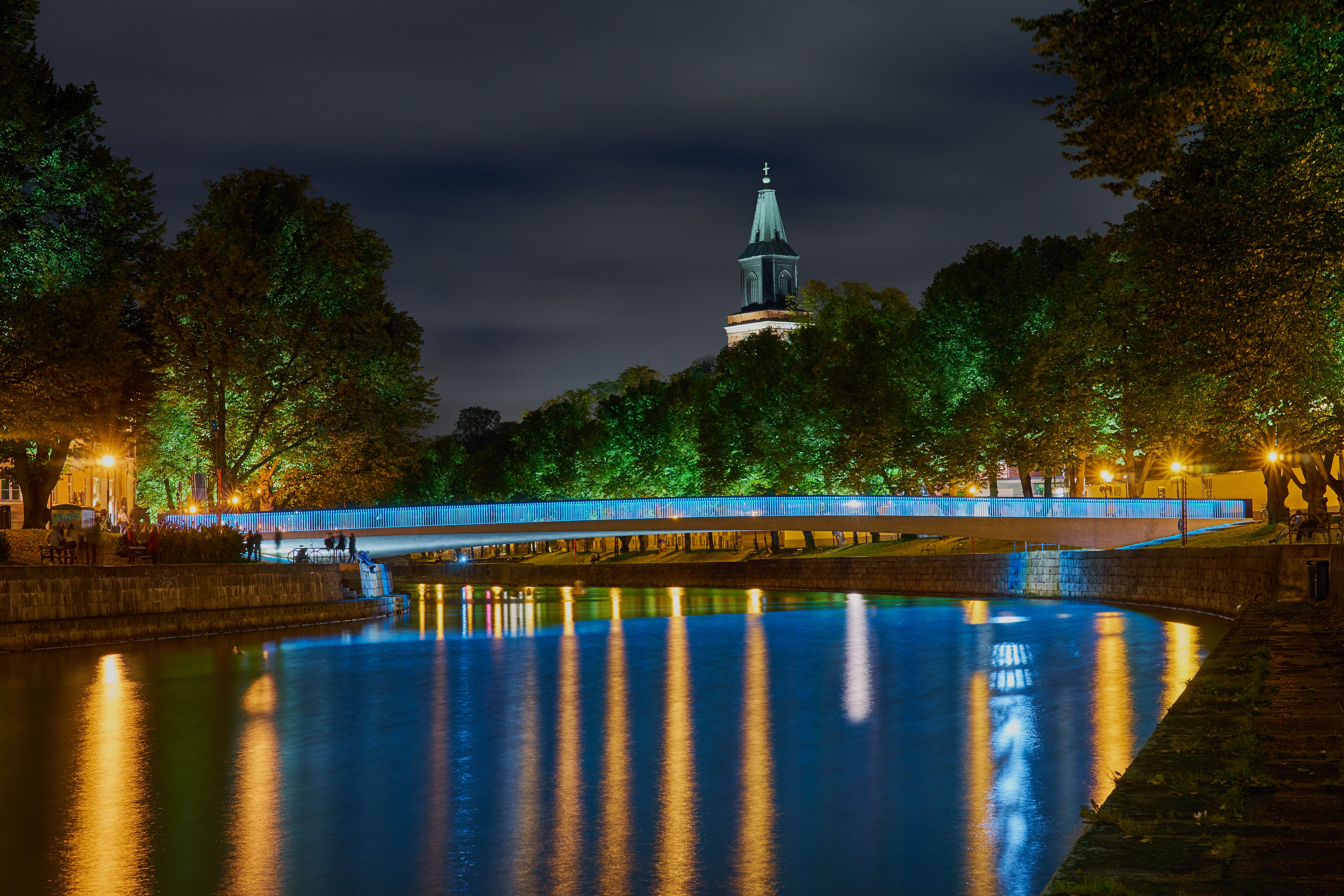 En natteudsigt over en flod med en blåoplyst bro, oplyste træer, et kirkespir og refleksioner i vandet.