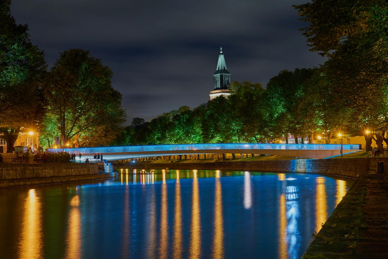 En natteudsigt over en flod med en blåoplyst bro, oplyste træer, et kirkespir og refleksioner i vandet.