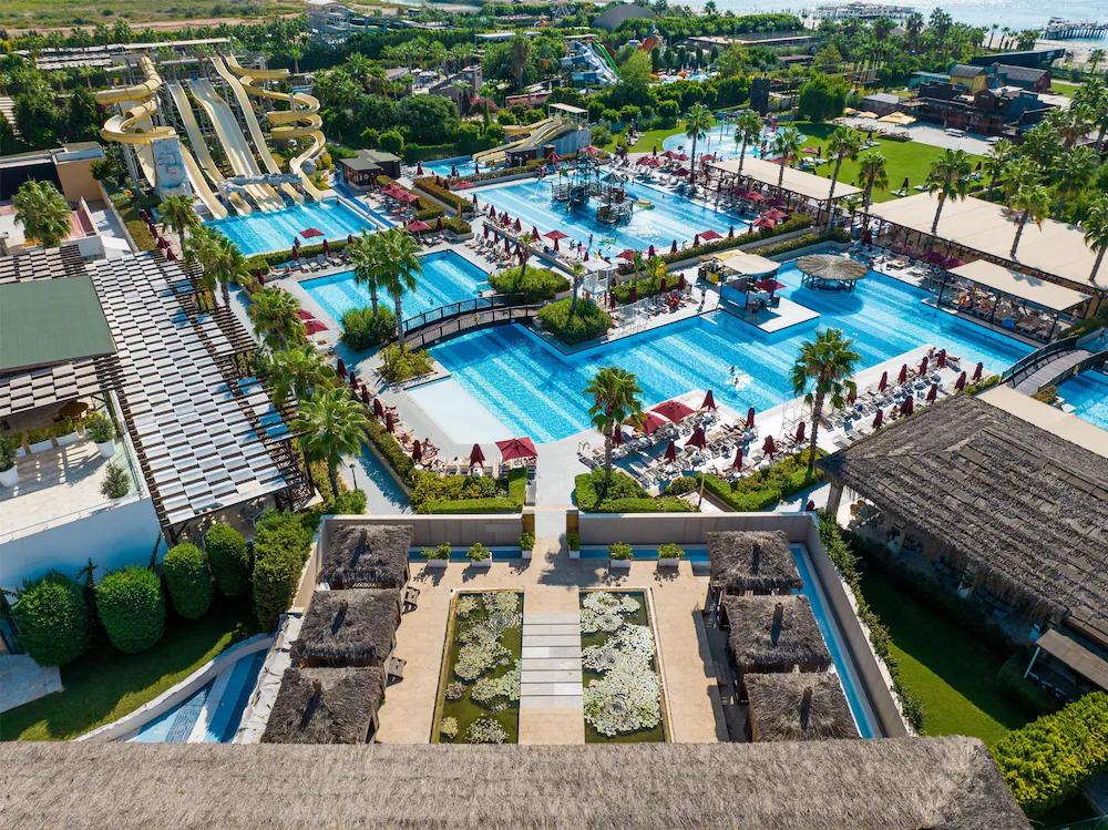 Luftfoto af en livlig resort med flere pools, vandrutsjebaner, palmer, liggestole og stråtækte bygninger, med havet i det fjerne. Aska Lara Resort & Spa