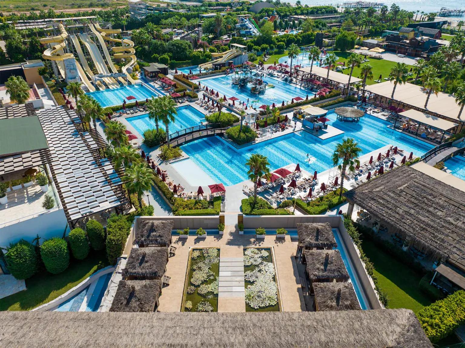 Luftfoto af en livlig resort med flere pools, vandrutsjebaner, palmer, liggestole og stråtækte bygninger, med havet i det fjerne. Aska Lara Resort & Spa