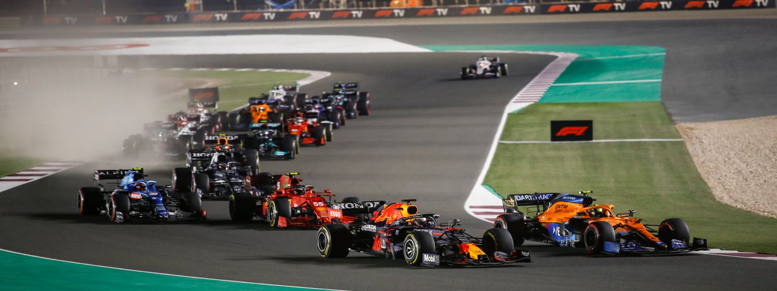 A pack of Formula 1 cars racing at night on a track.