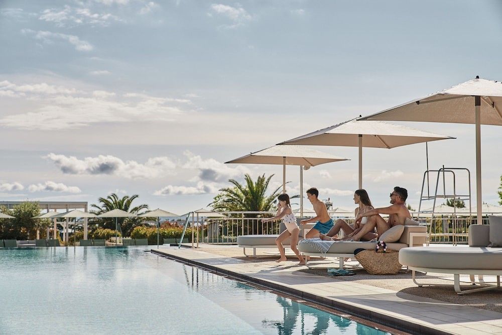 En familie på fire, der nyder et stort udendørs poolområde med parasoller og liggestole. Falkensteiner Family Hotel Diadora