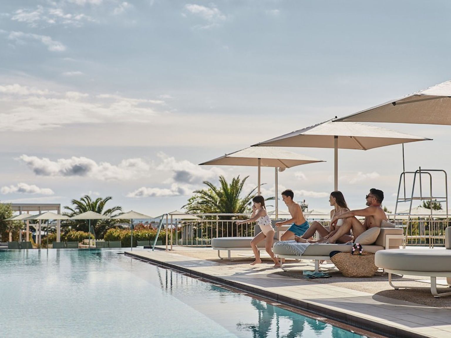 En familie på fire, der nyder et stort udendørs poolområde med parasoller og liggestole. Falkensteiner Family Hotel Diadora