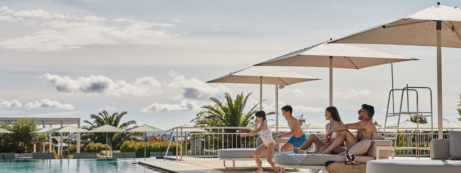 En familie på fire, der nyder et stort udendørs poolområde med parasoller og liggestole. Falkensteiner Family Hotel Diadora