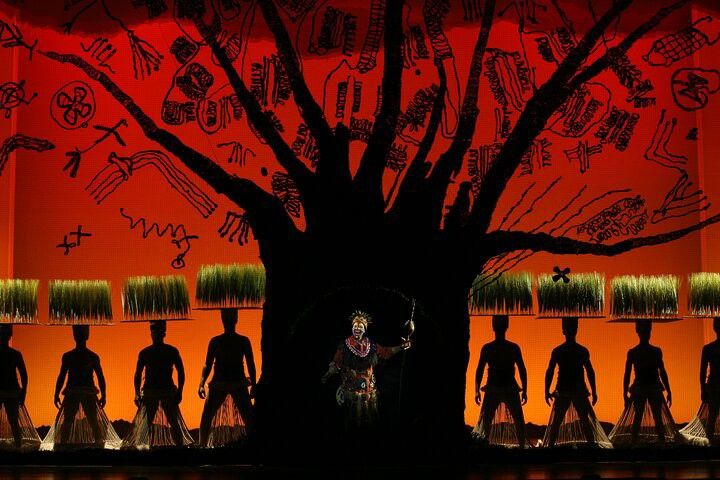 The Lion King musical: Rafiki, silhouetted performers with grass on their heads, and a dark tree against an orange, tribal-inspired backdrop.