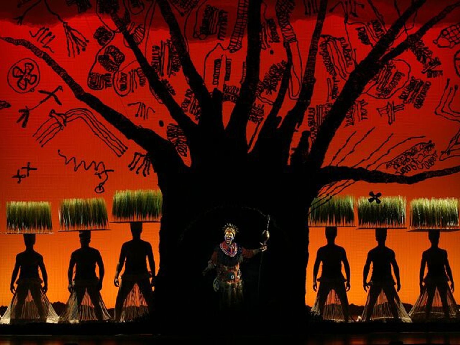 The Lion King musical: Rafiki, silhouetted performers with grass on their heads, and a dark tree against an orange, tribal-inspired backdrop.