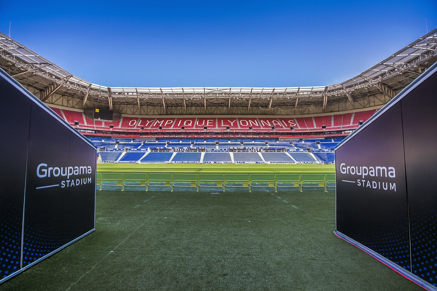 Blick vom Spielfeld-Eingang des Groupama-Stadions, mit "Groupama Stadium"-Schildern und "OLYMPIQUE LYONNAIS" in den leeren roten und blauen Sitzen ausgeschrieben.