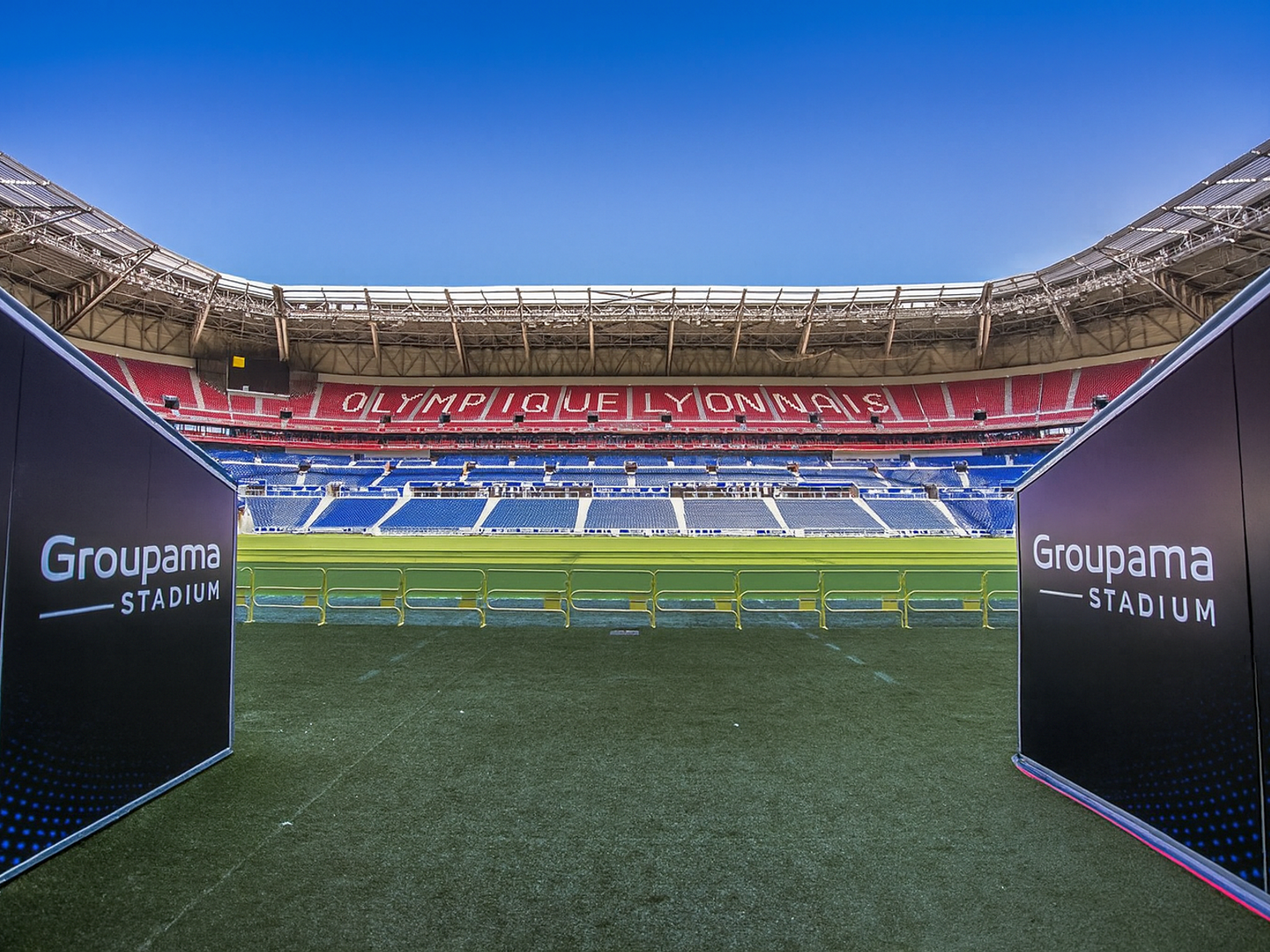 Blick vom Spielfeld-Eingang des Groupama-Stadions, mit "Groupama Stadium"-Schildern und "OLYMPIQUE LYONNAIS" in den leeren roten und blauen Sitzen ausgeschrieben.
