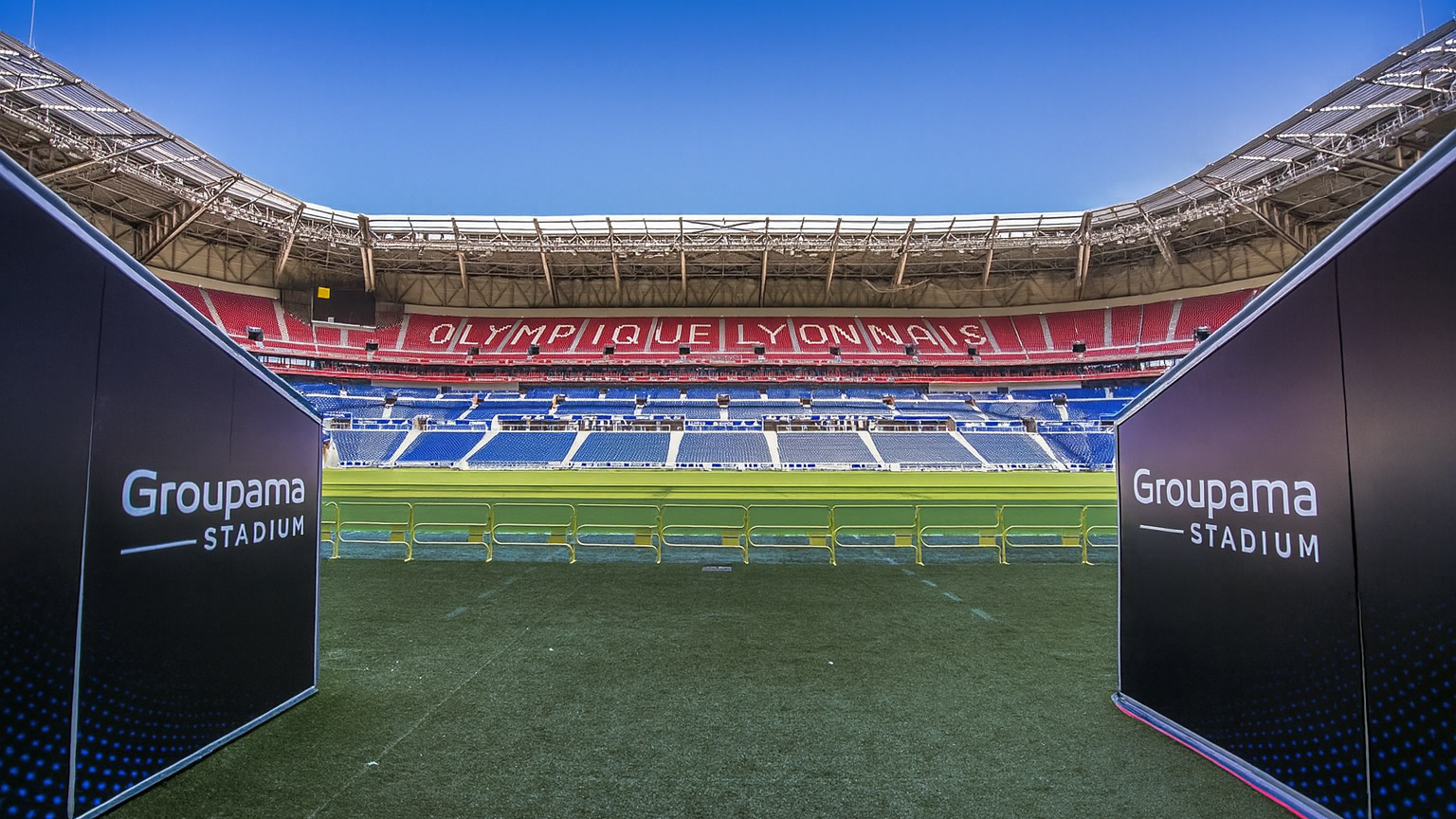 View from the pitch entrance of Groupama Stadium, with "Groupama Stadium" signs and "OLYMPIQUE LYONNAIS" spelled out in the empty red and blue seats.