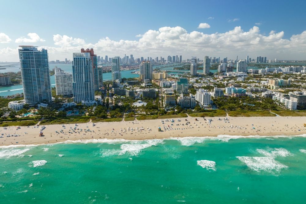 Luftfoto af en solrig strand med turkis bølger, mange mennesker og en bysilhuet med høje bygninger. Miami Beach