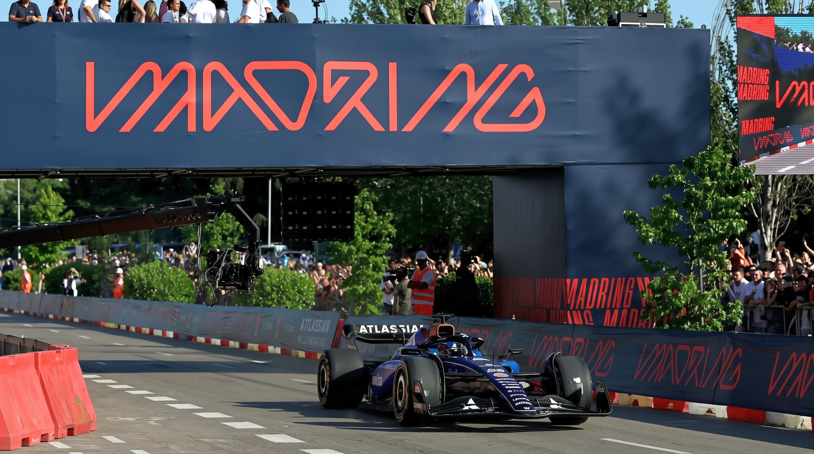 Une voiture bleue de style Formule 1 roule sur un circuit sous une banderole « MADRING », devant une foule de spectateurs.