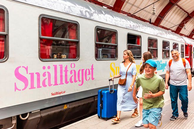 En familj, med en pojke som springer och en kvinna som drar en blå resväska, går på en tågplattform bredvid ett Snälltåget-tåg.