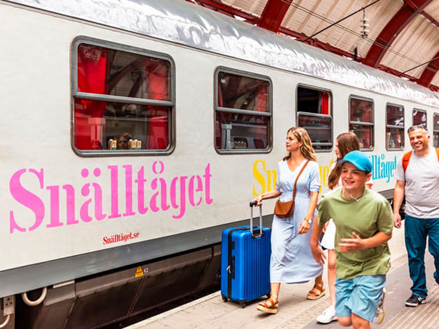 En familj, med en pojke som springer och en kvinna som drar en blå resväska, går på en tågplattform bredvid ett Snälltåget-tåg.