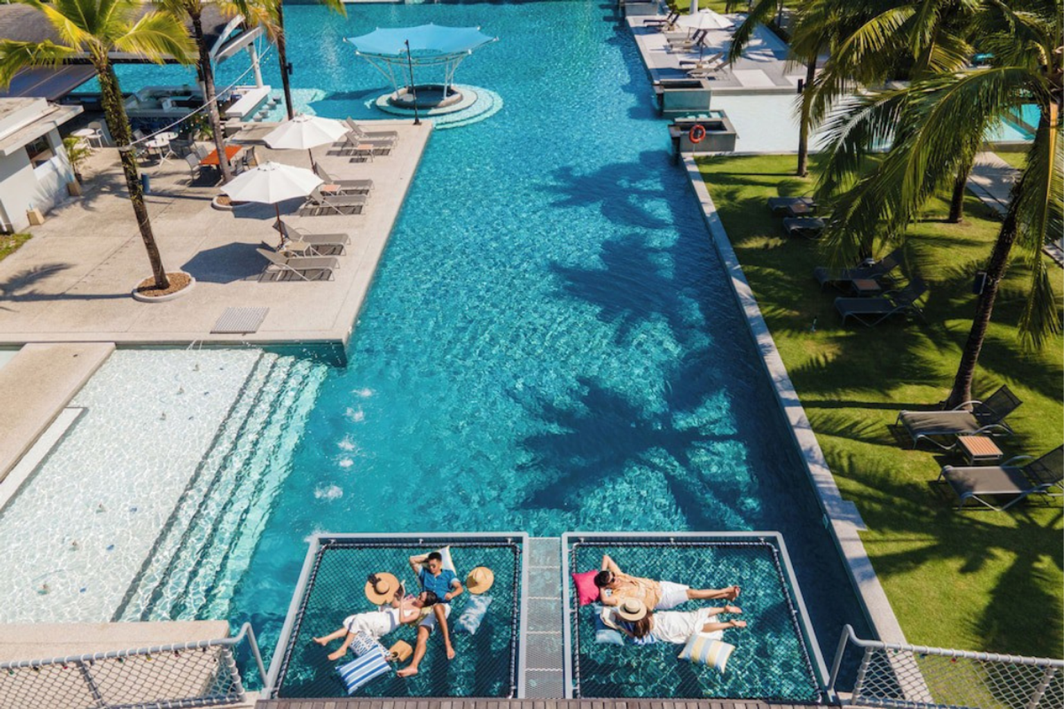 Flygvy över ett poolområde på en resort med människor som kopplar av i nedsänkta hängmattor, palmer och solstolar.