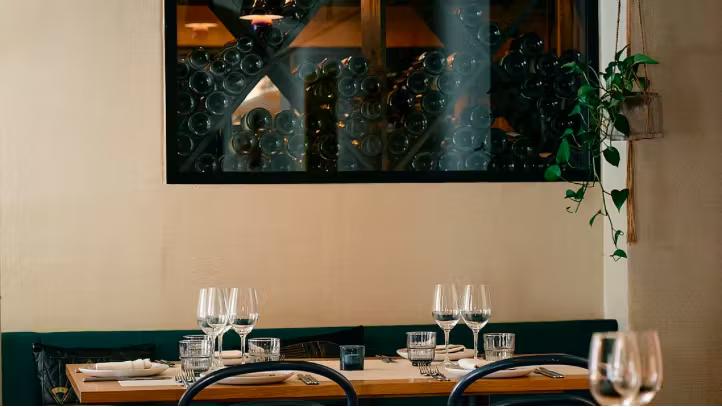 Et restaurantbord dækket med glas og tallerkener, en mørkegrøn bås, en dekorativ væg med vinflasker og en hængende plante.