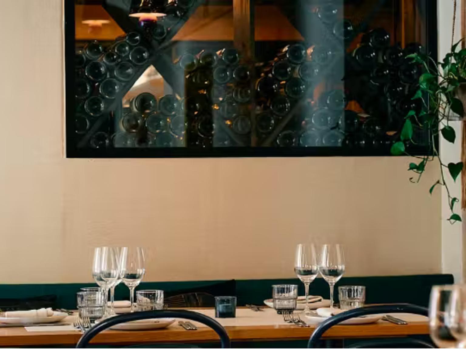 Et restaurantbord dækket med glas og tallerkener, en mørkegrøn bås, en dekorativ væg med vinflasker og en hængende plante.