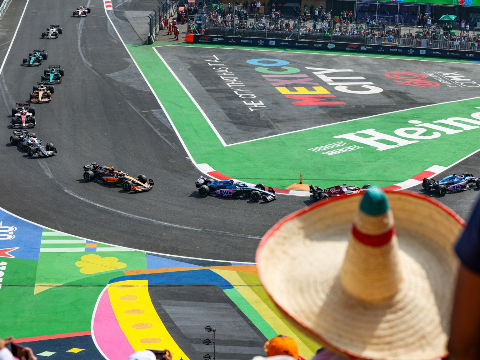 Formel 1-biler på en bane med "Mexico City" og "Heineken" logoer, sett over en tilskuerens sombrero.