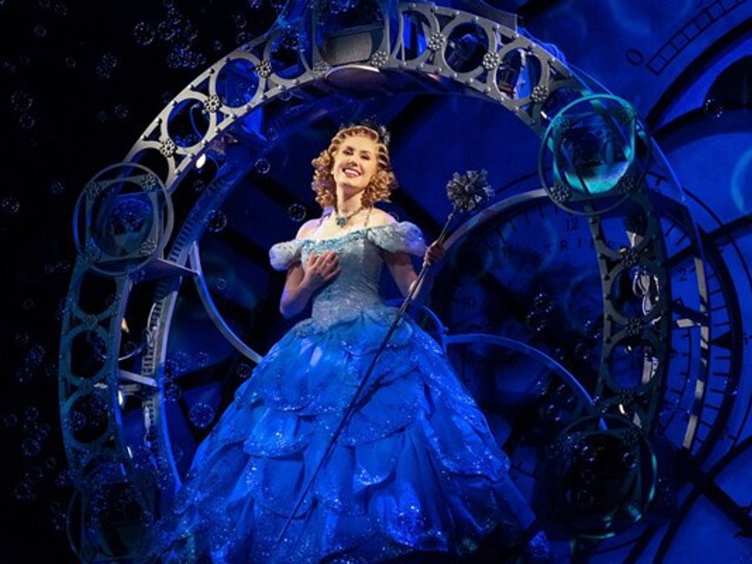 Glinda the Good Witch in a shimmering blue gown, holding a wand, within an ornate archway with bubbles.