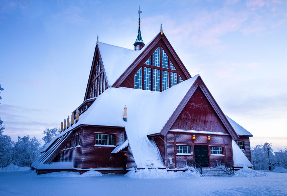 En mörkröd träkyrka med branta, snötäckta tak mot en blek vinterhimmel. Kiruna, Sverige