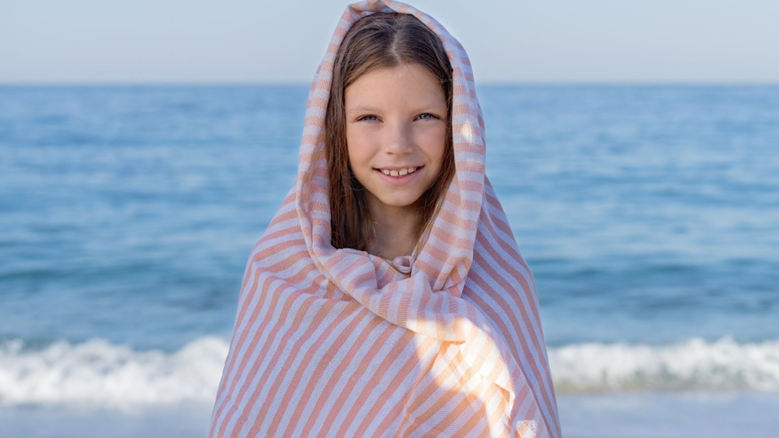 En smilende jente innhyllet i et fersken- og hvitstripet håndkle på stranden.