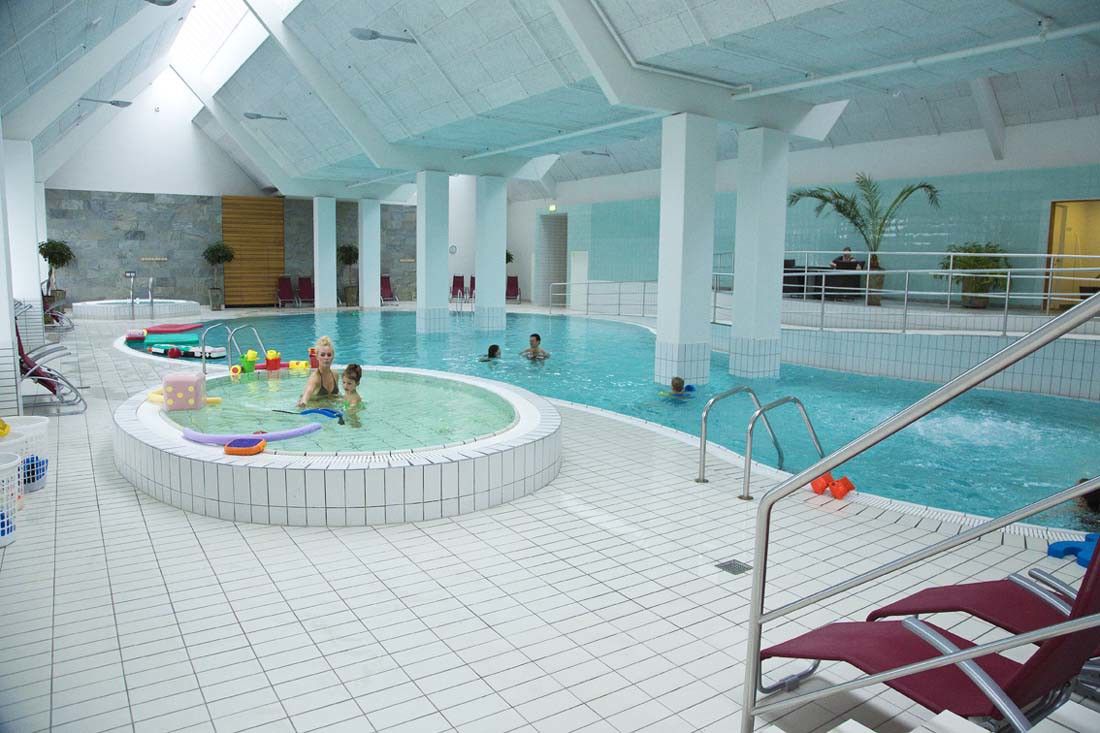 Ett ljust inomhusbad med en huvudpool, en grund barnpool där en kvinna och ett barn leker med leksaker, samt en jacuzzi.