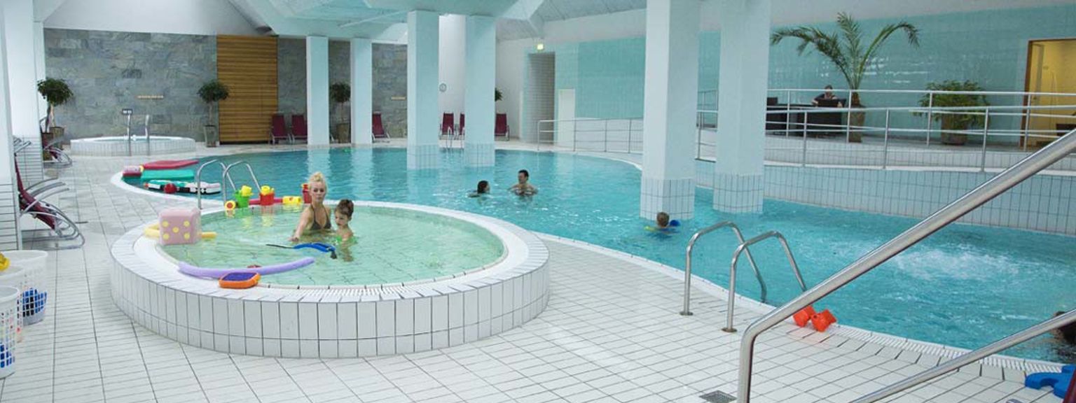 Ett ljust inomhusbad med en huvudpool, en grund barnpool där en kvinna och ett barn leker med leksaker, samt en jacuzzi.
