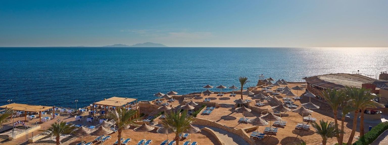Solig resortstrand med stråparasoller, solstolar och palmer med utsikt över det blå havet.