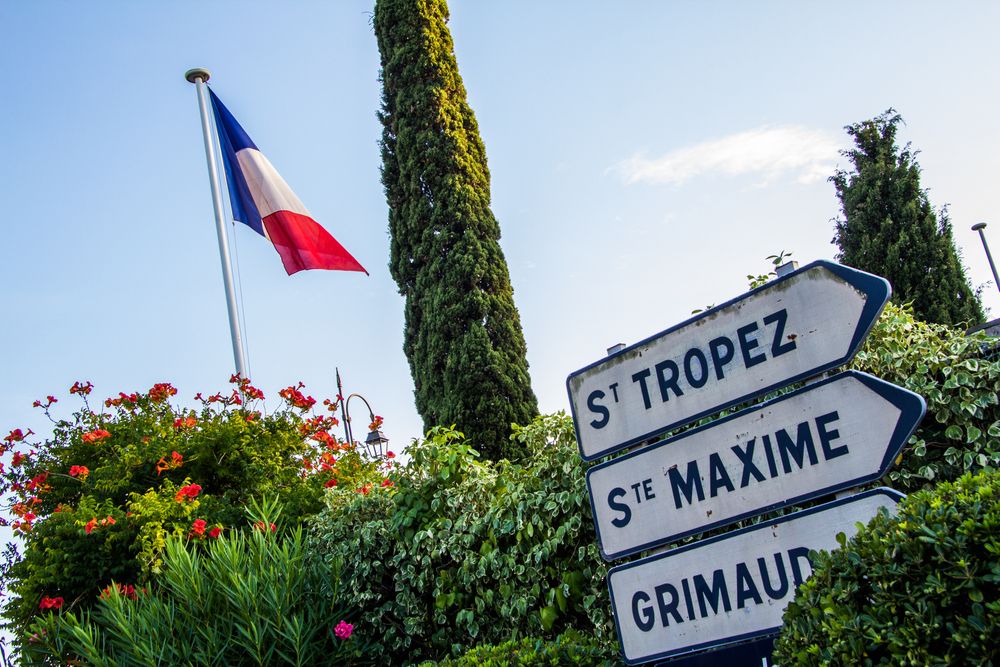 French flag flying above lush greenery and a signpost pointing to St. Tropez, Ste. Maxime, and Grimaud. Sainte-Maxime, France