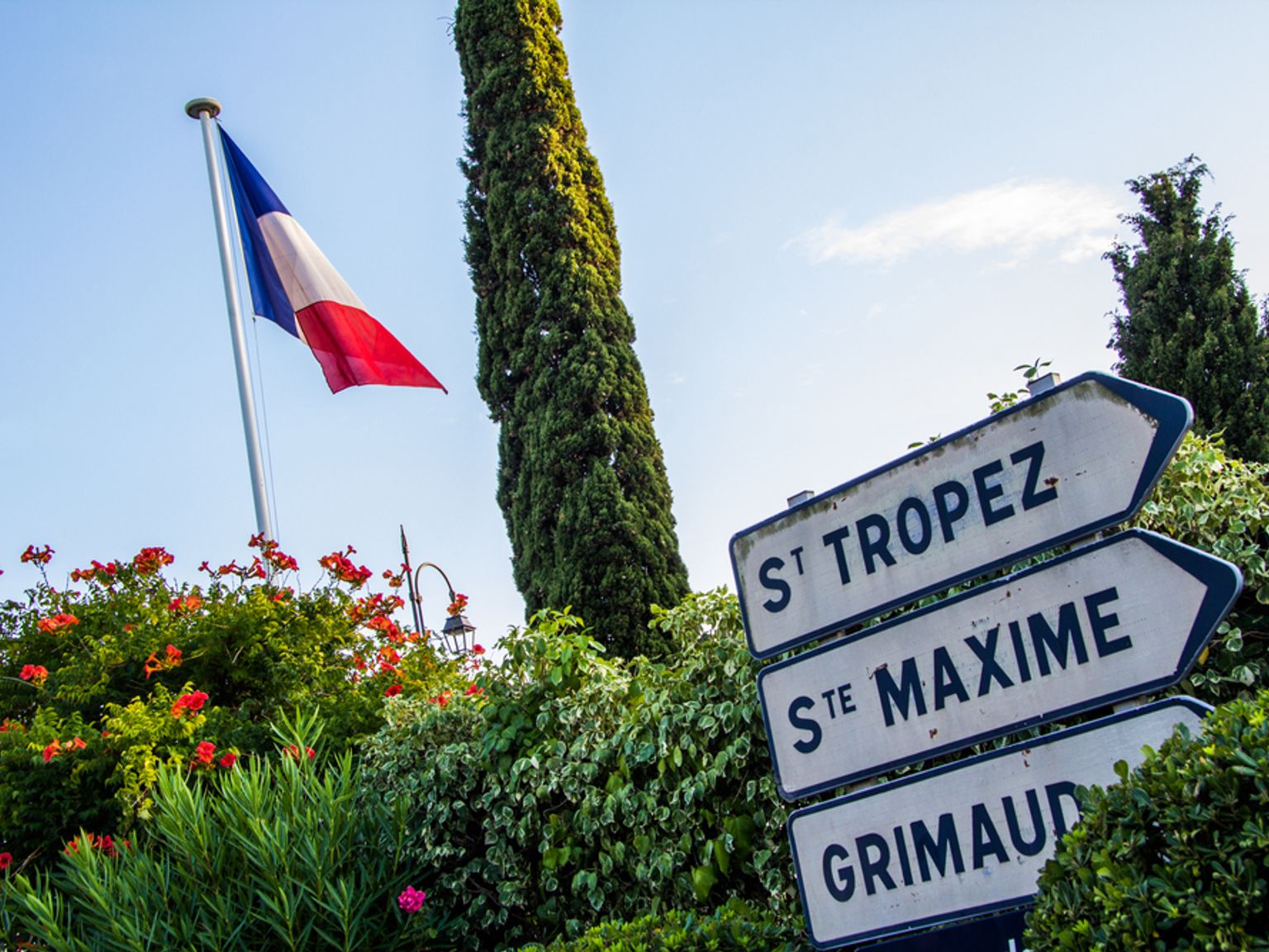 Drapeau français flottant au-dessus d’une végétation luxuriante et un panneau indicateur pointant vers St. Tropez, Ste. Maxime et Grimaud. Sainte-Maxime, France